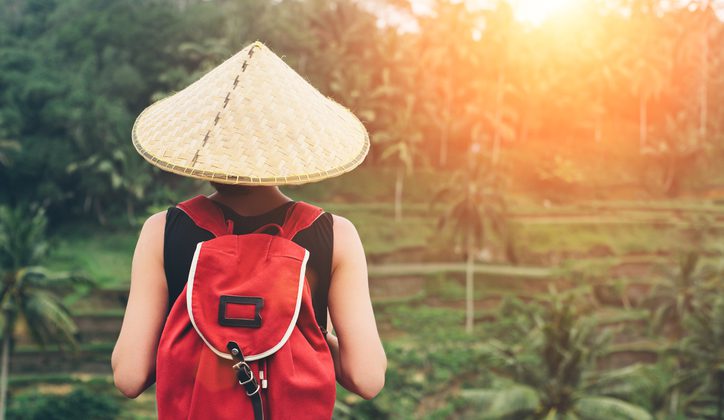 Young lady with traditional Asian hat and backpack standing and looking at tea plants (intentional sun glare)